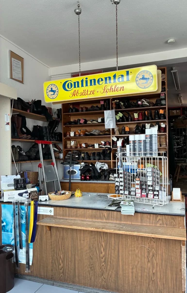 Verkaufsraum der Schuhmacherei: Holztheke, Regale voller Schuhe und darüber das gelbe Continental-Schild „Absätze Sohlen“
